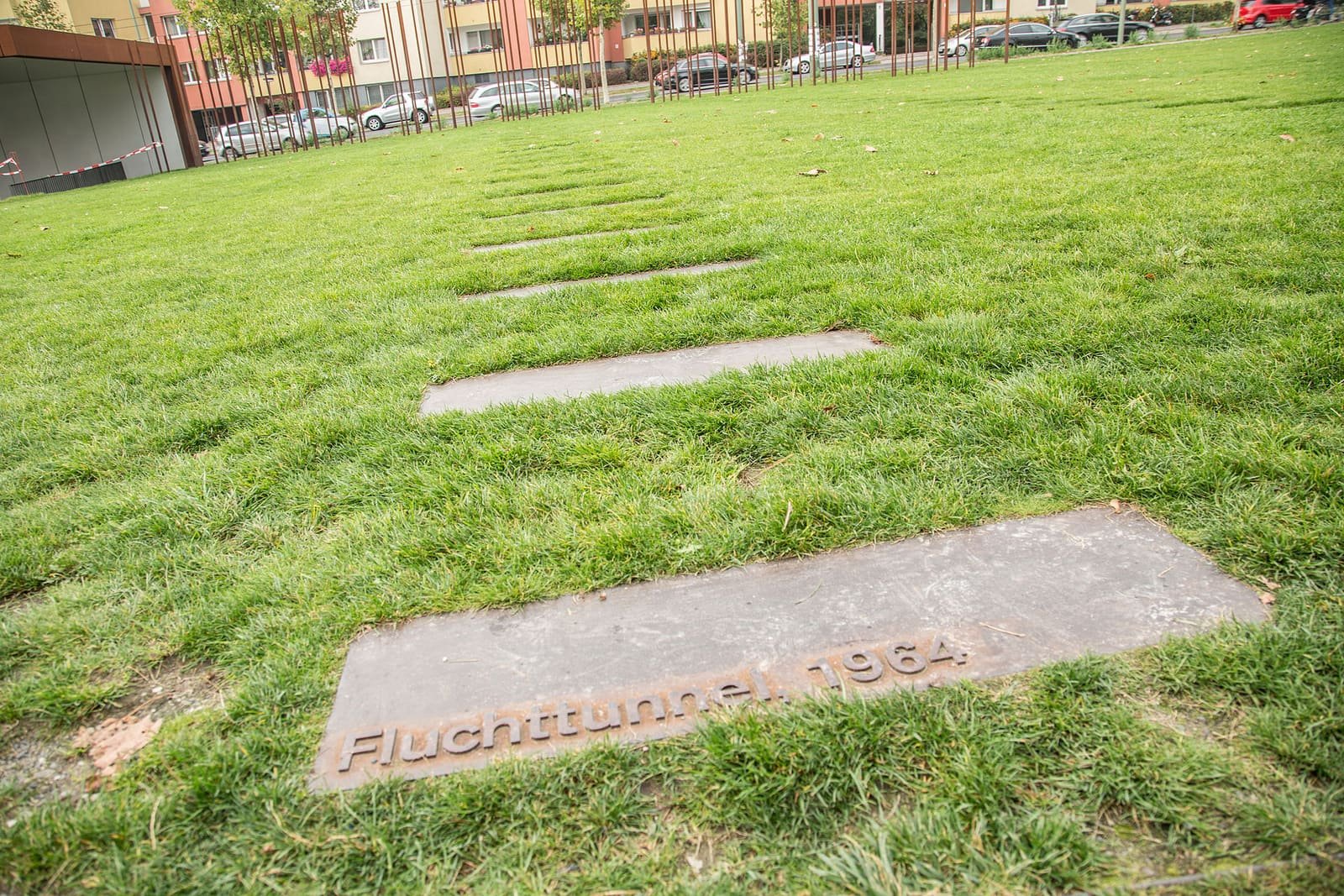 A lawn with a line of rectangular steel plates set into the grass; the nearest plate is inscribed 'Fluchttunnel 1964'. A row of rusty steel rods marking the Wall's course and apartment buildings are in the background.