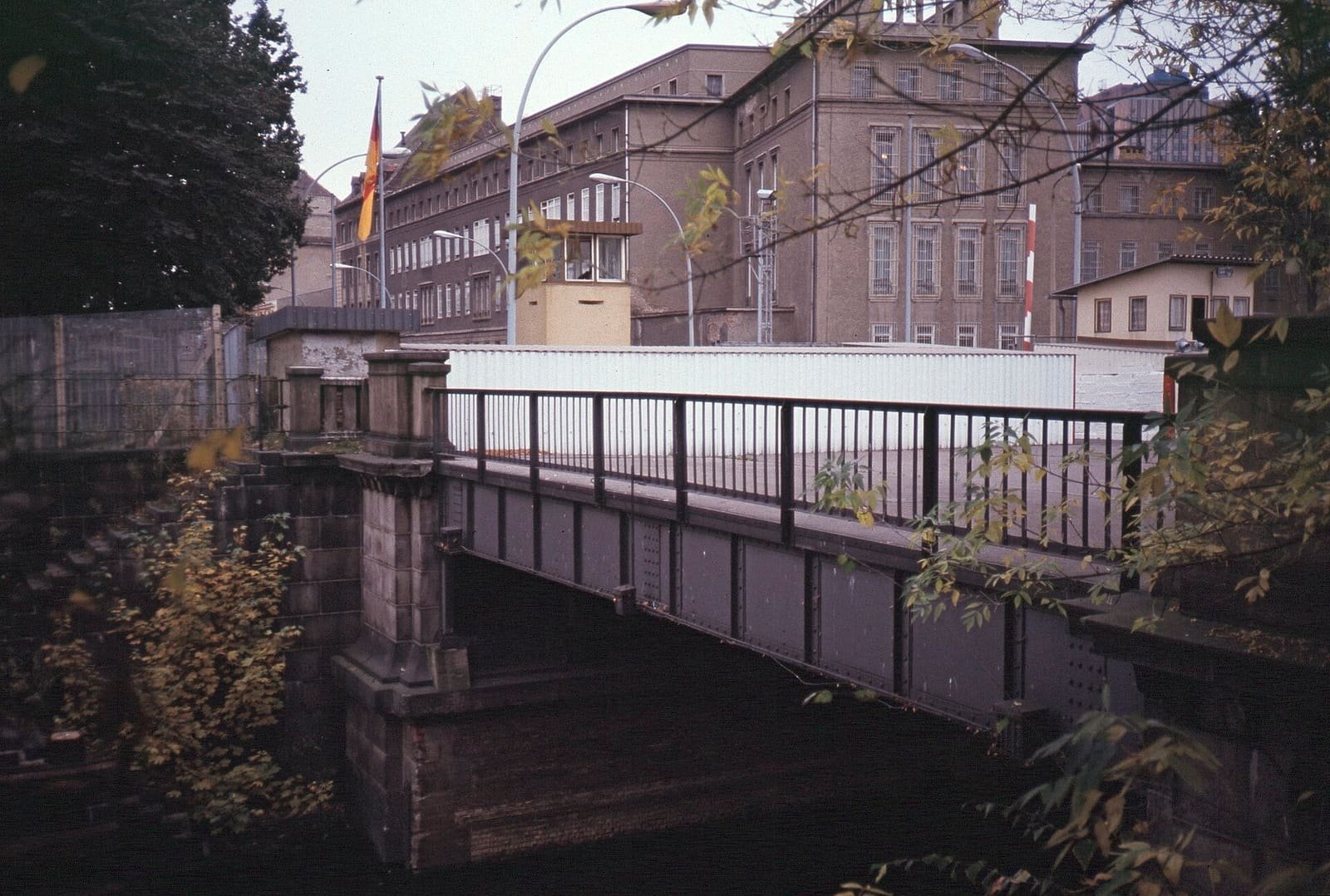 Colour photo of a small iron road bridge over a canal leading to a white barrier wall, a guard booth and large grey administrative buildings, framed by autumn leaves.
