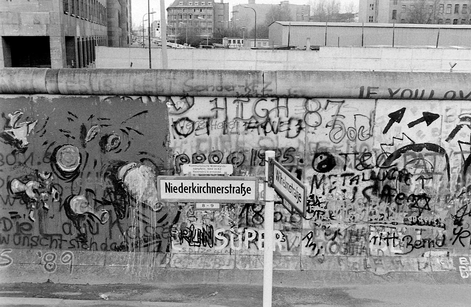 Black-and-white photo: street signs for Niederkirchnerstraße and Wilhelmstraße in front of a graffiti-covered wall; behind it an empty strip, a second wall and large stone buildings.